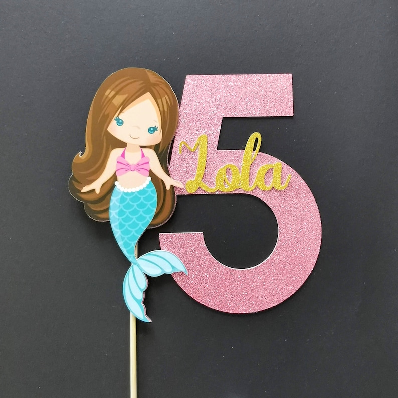 Puede incluir: Topper de pastel de n&uacute;mero cinco de purpurina rosa con una ilustraci&oacute;n de sirena y el nombre "Lola" en purpurina dorada.