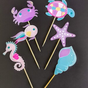 Puede incluir: Seis adornos para pasteles de criaturas marinas de colores en palos de madera. Los adornos incluyen un cangrejo morado, un caballito de mar rosa, una concha azul, una estrella de mar rosa y azul, una tortuga rosa y azul y un pez rosa y amarillo.