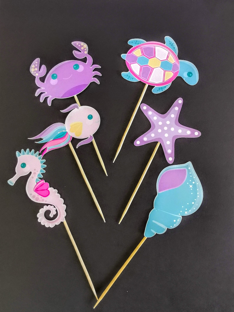 Puede incluir: Seis adornos para pasteles de criaturas marinas de colores en palos de madera. Los adornos incluyen un cangrejo morado, un caballito de mar rosa, una concha azul, una estrella de mar rosa y azul, una tortuga rosa y azul y un pez rosa y amarillo.