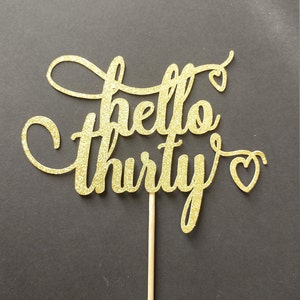 May include: A gold glitter "hello thirty" cake topper with a wooden stick. The script font has decorative swirls and heart accents. The topper is set against a dark background.