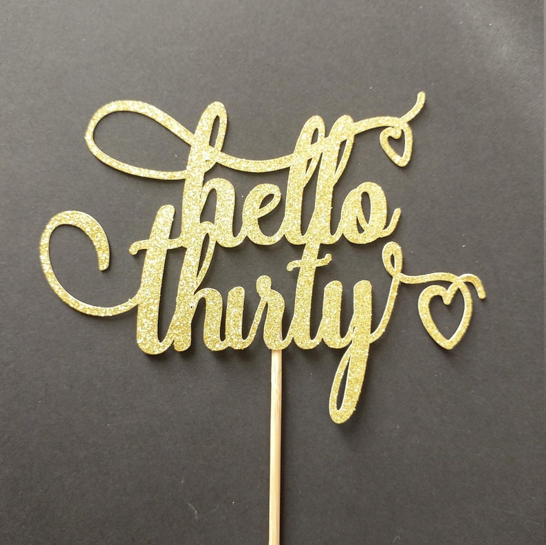 May include: A gold glitter "hello thirty" cake topper with a wooden stick. The script font has decorative swirls and heart accents. The topper is set against a dark background.