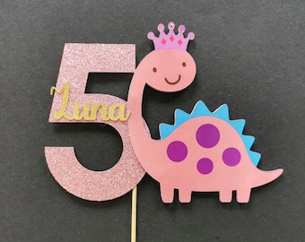 May include: Pink glitter number 5 cake topper with the name "Luna" written in gold glitter. A pink dinosaur with a blue back and purple spots wears a pink crown and smiles.