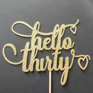 May include: A gold glitter cake topper with the words "hello thirty" in a cursive font. The word "hello" is above the word "thirty". Small heart shapes are at the end of the words. The topper is attached to a wooden stick.
