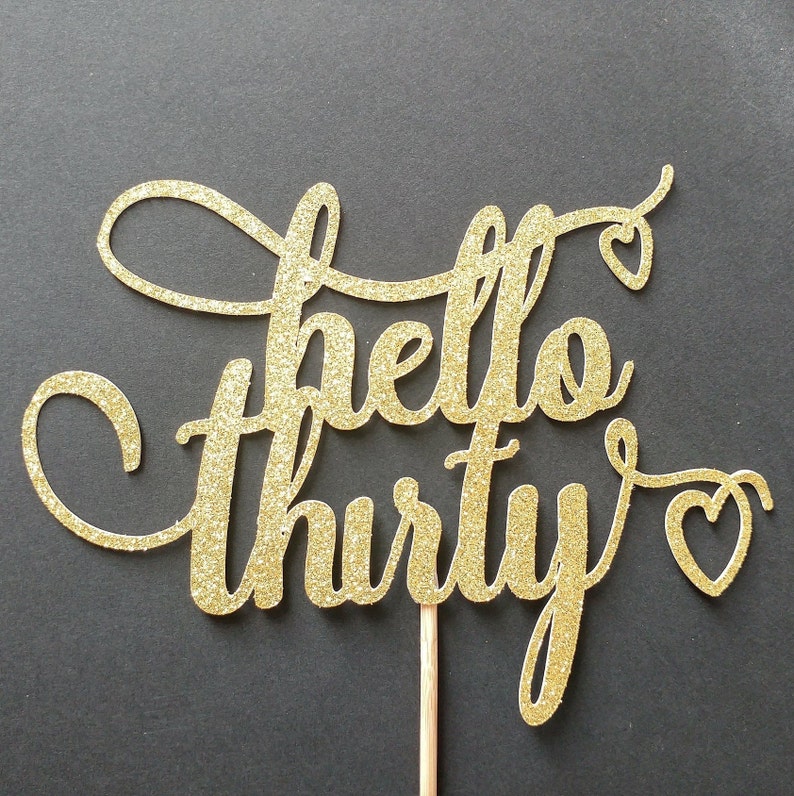 May include: A gold glitter cake topper with the words "hello thirty" in a cursive font. The word "hello" is above the word "thirty". Small heart shapes are at the end of the words. The topper is attached to a wooden stick.