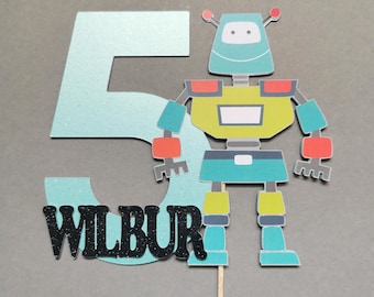 May include: A turquoise blue number five with glitter, the word "WILBUR" in black glitter, and a robot cake topper. The robot is green, yellow, blue, and red.