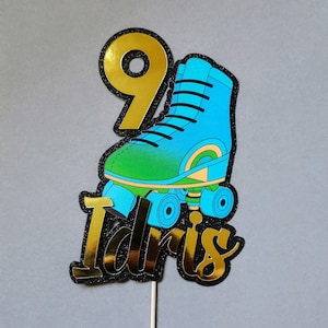 Peut inclure: Un topper de gâteau en paillettes dorées avec le chiffre 9 et le nom "Idris" en paillettes dorées. Le chiffre 9 est sur un patin à roulettes bleu et vert avec des roues noires.