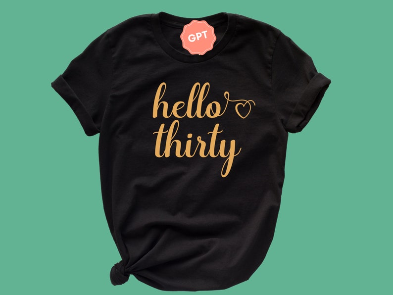 May include: Black t-shirt with the words "hello thirty" in a gold script font. A small heart graphic is above the word "thirty". The t-shirt is knotted at the bottom and has rolled-up sleeves.