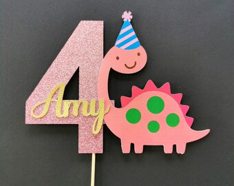 May include: Pink glitter number four cake topper with a pink dinosaur wearing a blue and white striped party hat. The dinosaur has green spots and the number four has the name "Amy" written in gold glitter.