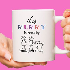 K&ouml;nnte beinhalten: Wei&szlig;e Keramiktasse mit einer Schwarzwei&szlig;illustration einer Familie mit einer Katze. Der Text lautet "this MUMMY is loved by Emily Josh Cindy".
