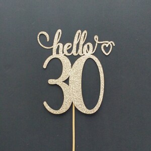 May include: Gold glitter cake topper with the text "hello" and a heart, and the number "30" in a cursive font.