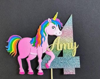 May include: A pink unicorn cake topper with a rainbow mane and tail, a gold horn, and the name "Amy" in gold script. The number "4" is glittery with pink, blue, and silver hues. The topper is attached to a wooden stick.