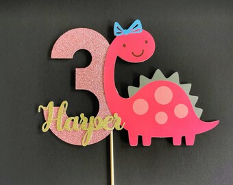 May include: Pink glitter number three cake topper with a pink dinosaur with a blue bow and grey spikes. The dinosaur has pink polka dots and the name "Harper" is written in gold glitter.