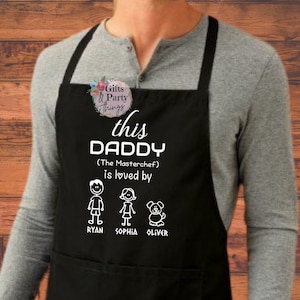 以下が含まれることがあります： 「this DADDY (The Masterchef) is loved by」のテキストと、男の子、女の子、犬の棒人間イラストが描かれた黒いエプロン。名前はRyan、Sophia、Oliver。エプロンにはフロントポケットが付いています。