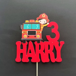 May include: Red and black fire truck cake topper with a firefighter holding a hose. The topper also includes the number 3 and the name Harry.