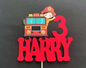 May include: Red and black fire truck cake topper with a firefighter holding a hose. The topper also includes the number 3 and the name Harry.