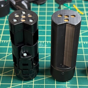 Puede incluir: Dos objetos cilíndricos negros con tapas metálicas. Uno tiene un diseño segmentado con tornillos visibles, mientras que el otro es un cilindro sólido con un acabado de metal cepillado. Ambos tienen aberturas circulares.