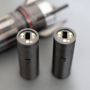 Puede incluir: Dos objetos cilíndricos de color gris oscuro con una abertura cuadrada en la parte superior y pequeños círculos dorados. Un objeto gris metálico está en el fondo, parcialmente desenfocado.