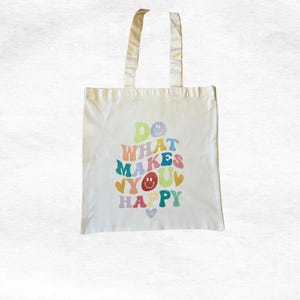 Op de afbeelding: Cr&egrave;mekleurige canvas draagtas met de tekst "DO WHAT MAKES YOU HAPPY" in kleurrijke retroletters. Het ontwerp bevat smileys en harten in verschillende kleuren, met bijpassende cr&egrave;mekleurige handvatten.
