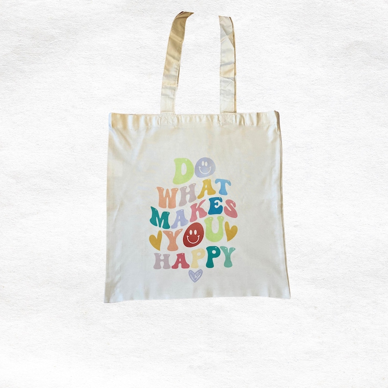 Op de afbeelding: Cr&egrave;mekleurige canvas draagtas met de tekst "DO WHAT MAKES YOU HAPPY" in kleurrijke retroletters. Het ontwerp bevat smileys en harten in verschillende kleuren, met bijpassende cr&egrave;mekleurige handvatten.