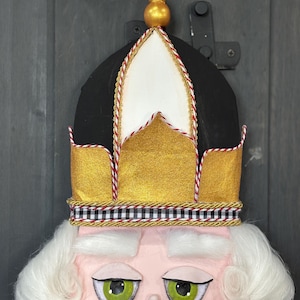 May include: A close-up of a decorative nutcracker head. The nutcracker has a black and gold crown with red and gold trim. The face is pink with green eyes, white hair, and a white mustache. The background is a dark gray wooden door.
