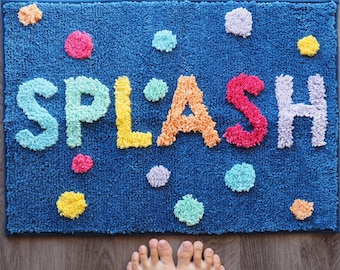 Puede incluir: Una alfombra de baño rectangular azul con textura de mechones que presenta la palabra SPLASH en letras grandes y multicolores. La alfombra está decorada con varios puntos circulares y multicolores dispersos por la superficie en tonos rosa, amarillo, naranja, azul claro y lavanda.