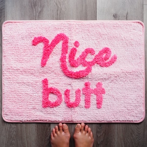 May include: Pink bath mat with the words "Nice but" in pink fluffy letters.