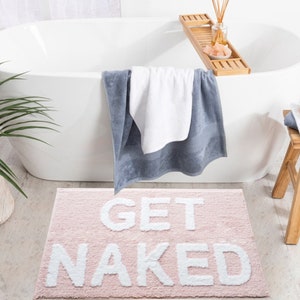 Puede incluir: Una alfombra de ba&ntilde;o rosa con las palabras "GET NAKED" en blanco, colocada frente a una ba&ntilde;era blanca. Una toalla azul y blanca cuelga de la ba&ntilde;era. Una bandeja de madera con accesorios de ba&ntilde;o est&aacute; en el borde de la ba&ntilde;era.