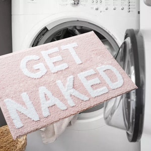Puede incluir: Una alfombra de ba&ntilde;o rectangular rosa con las palabras "GET NAKED" en letras blancas grandes. La alfombra est&aacute; hecha de un material suave y texturizado. La alfombra se sostiene frente a una lavadora.