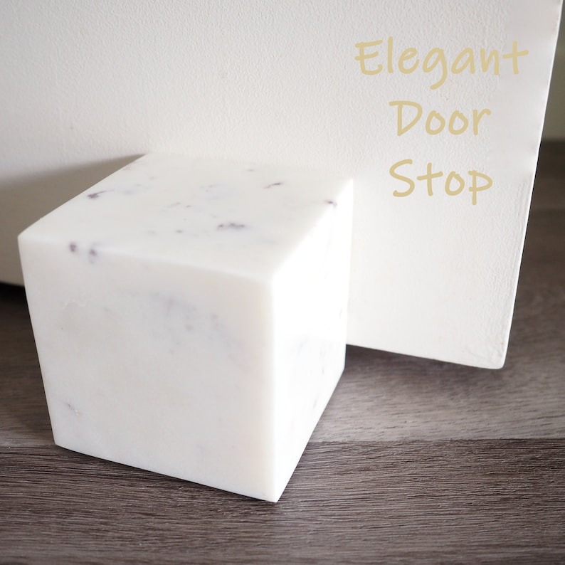 Op de afbeelding: Een witte marmeren deurstop met een gespikkeld patroon. De deurstop is kubusvormig en staat op een houten vloer. De tekst "Elegant Door Stop" staat op een wit oppervlak achter de deurstop.