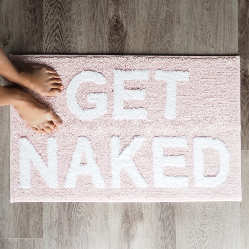 Puede incluir: Una alfombra de ba&ntilde;o rectangular de color rosa claro con las palabras "GET NAKED" en letras grandes, blancas y texturizadas. La alfombra est&aacute; sobre un suelo de madera, con los pies de una persona visibles en la parte superior.