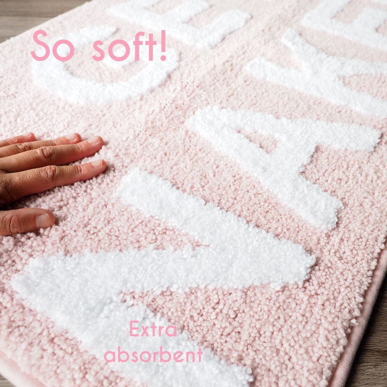 Puede incluir: Una alfombra de ba&ntilde;o rosa con letras blancas en relieve que deletrean un nombre. La alfombra es suave y absorbente, con las palabras "So soft!" y "Extra absorbent" en texto rosa.