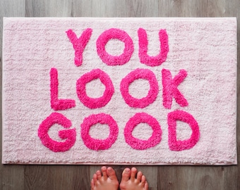 May include: A rectangular pink bath mat with the words "YOU LOOK GOOD" in bright pink, fluffy letters. The mat is on a wooden floor, and a pair of feet are visible at the bottom of the image.