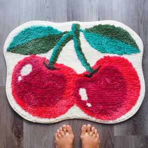 May include: A white bath mat with a red and green cherry design. The cherries are fuzzy and have a 3D effect.