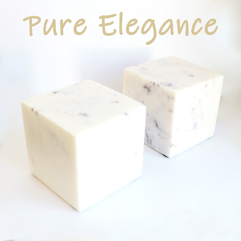 Op de afbeelding: Twee witte marmeren kubussen met een gespikkeld patroon. De kubussen staan op een wit oppervlak. De tekst "Pure Elegance" is in goud boven de kubussen geschreven.