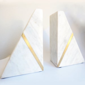 May include: A pair of white marble bookends with gold accents. Each bookend is shaped like a triangle with a gold stripe running diagonally across the front.