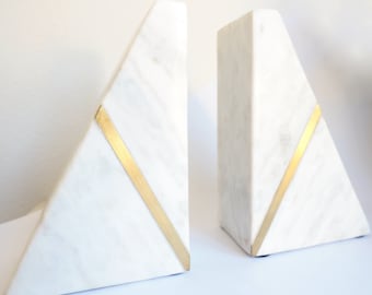 May include: A pair of white marble bookends with gold accents. Each bookend is shaped like a triangle with a gold stripe running diagonally across the front.