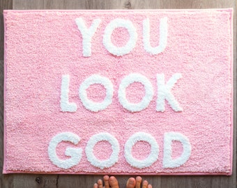 May include: Pink bath mat with the words "YOU LOOK GOOD" in white fluffy letters.