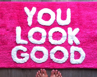 May include: A bright pink bath mat with the words "YOU LOOK GOOD" in white, fluffy letters. The mat is rectangular and has a textured surface. Bare feet are visible at the bottom of the mat.
