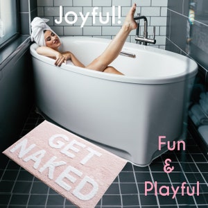 Puede incluir: Una alfombra de ba&ntilde;o rosa con las palabras "GET NAKED" en blanco, colocada cerca de una ba&ntilde;era blanca. La imagen tambi&eacute;n incluye las palabras "Joyful!" y "Fun & Playful".