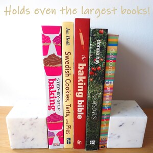 Op de afbeelding: Witte marmeren boekenleggers met een gevlekt patroon, die een rij kleurrijke kookboeken vasthouden. De boeken bevatten titels als "The Baking Bible", "Swedish Cookies, Tarts, and Pies" en "Delicious. 5 of the Best".