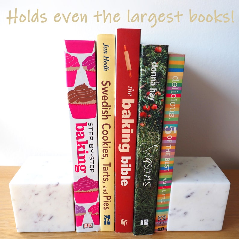 Op de afbeelding: Witte marmeren boekenleggers met een gevlekt patroon, die een rij kleurrijke kookboeken vasthouden. De boeken bevatten titels als "The Baking Bible", "Swedish Cookies, Tarts, and Pies" en "Delicious. 5 of the Best".