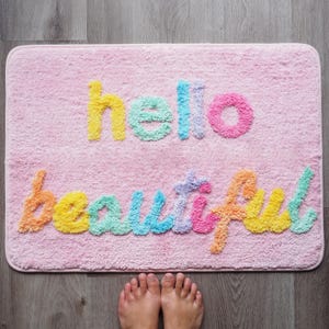 May include: A pink bath mat with the words "hello beautiful" in colorful, textured lettering. The letters are yellow, blue, pink, orange, and green. The mat is rectangular and has rounded corners.