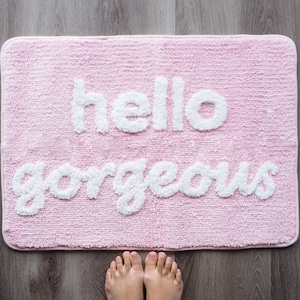 May include: A rectangular pink bath mat with the words "hello gorgeous" in white, fluffy lettering. The mat is on a wooden floor, and two feet are visible at the bottom of the image.