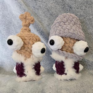 Puede incluir: Dos criaturas hechas a mano con ganchillo y grandes ojos saltones. Una es de color beige con una parte superior en forma de estrella, la otra es gris. Ambas tienen detalles en blanco y burdeos y pelaje blanco y esponjoso en la base. Son caprichosas y únicas.