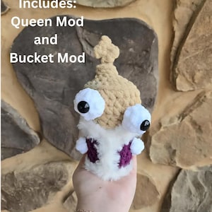 May include: A crocheted plush toy featuring a tan top section with a cross-shaped finial, two large white eyes with black pupils, and a white body with dark purple accents. The text "Includes: Queen Mod and Bucket Mod" is visible on the image.