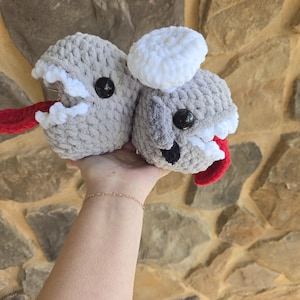 May include: Two handmade crochet creatures in gray with white and red accents. Each has a black button eye and a red tongue. One has a white fluffy top. The creatures are held up against a stone background.