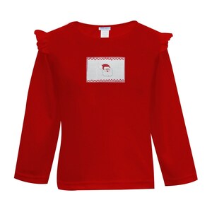May include: A bright red long-sleeved shirt with ruffle detailing at the shoulders. The shirt features a rectangular patch with a Santa Claus design embroidered in red and white. A festive holiday-themed garment.