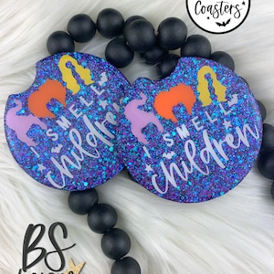 May include: Two glittery car coasters with a purple and blue background. The coasters feature the phrase "I SMELL CHILDREN" in white, with silhouettes of witches' hair in orange, yellow, and purple. The coasters are decorated with bats and stars.