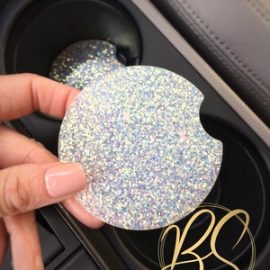 May include: A white glitter car coaster with a holographic finish. The coaster is round and has a slight curve to it. The coaster is being held in a hand and is being placed into a car cup holder.