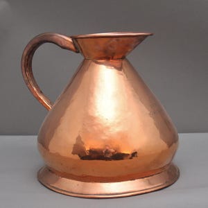May include: A copper pitcher with a wide base, a conical body, and a curved handle. The pitcher has a hammered texture and a spout. The surface reflects light, creating highlights and shadows. The pitcher is likely used for serving beverages.